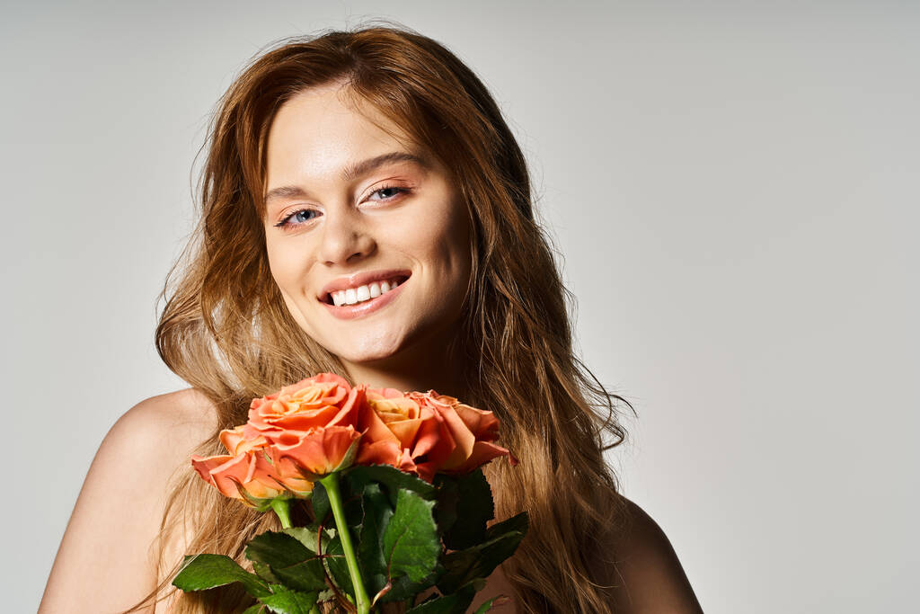 Portrait of smiling young woman with blue eyes, holding peachy roses posing on grey background - Photo, Image