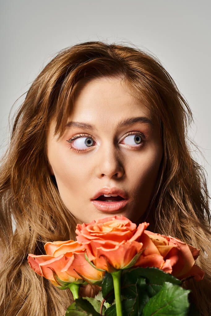 Портрет удивленной женщины с голубыми глазами, держащей персиковые розы, позирующие на сером фоне - Фото, изображение
