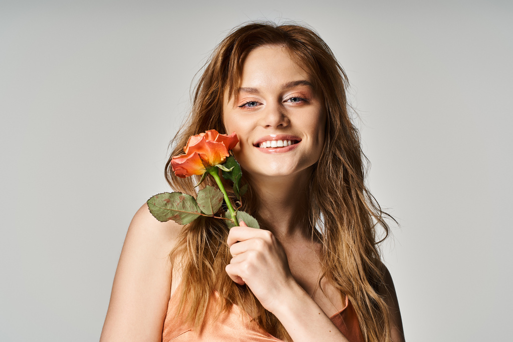 Χαρούμενο χαμογελαστό ελκυστικό κορίτσι με μπλε μάτια, με όμορφο τριαντάφυλλο κοντά στο πρόσωπο σε γκρι φόντο - Φωτογραφία, εικόνα