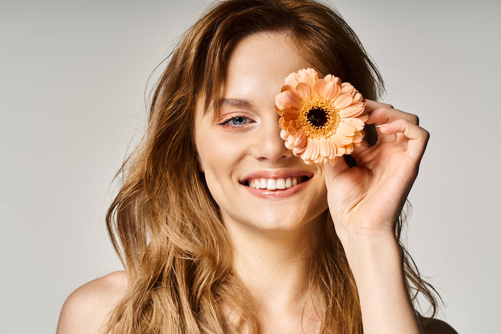 Piękna uśmiechnięta młoda kobieta z niebieskimi oczami, z gerbera daisy w pobliżu oka na szarym tle - Zdjęcie, obraz