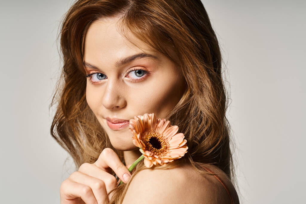 Side view of smiling attractive blue eyed girl, with gerbera daisy near face on grey background - Photo, Image