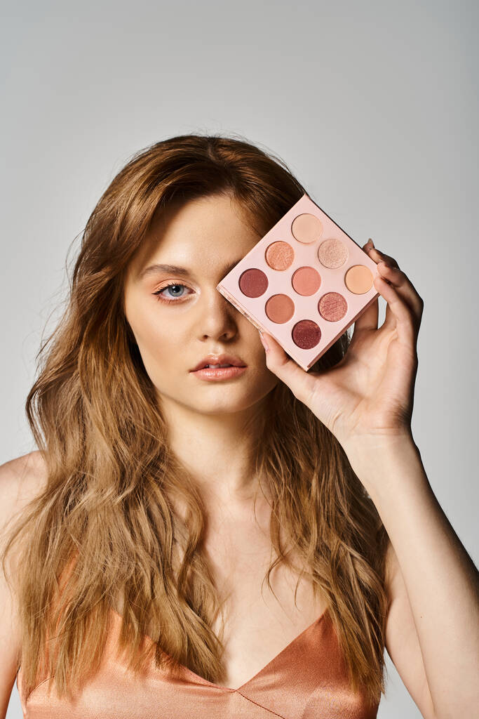 Beauty shot of woman holding peach makeup palette near eye on grey studio background - Photo, Image