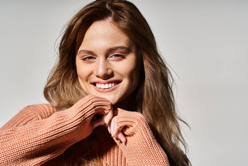 Студийный снимок улыбающейся женщины, смотрящей на камеру с натуральным макияжем, носящей свитер, трогающей подбородок - Фото, изображение
