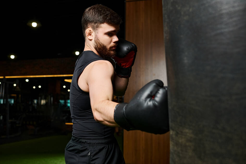 Un hombre elegante con barba, con una camiseta negra y guantes de boxeo, es visto golpeando una bolsa en un gimnasio. - Foto, Imagen