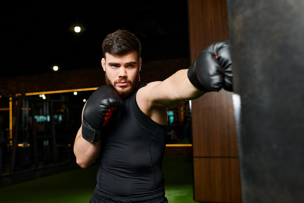 A bearded man in a black shirt trains in a gym, throwing punches at a heavy bag with black boxing gloves. - Photo, Image