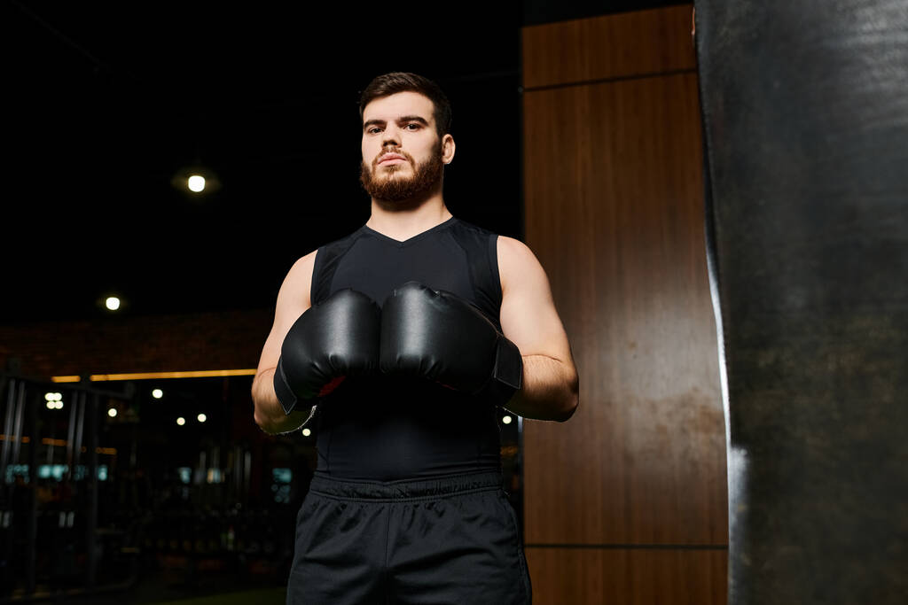 Egy szakállas, jóképű férfi bokszkesztyűben magabiztosan áll egy bokszzsák mellett egy edzőteremben.. - Fotó, kép