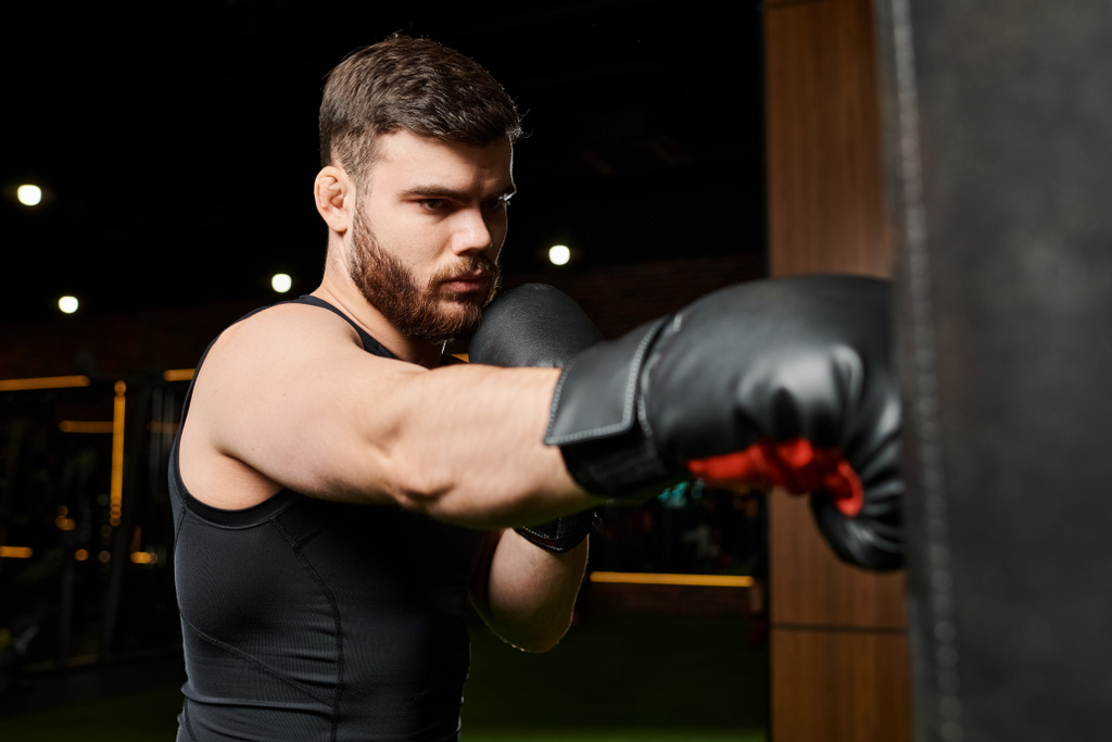 Un hombre guapo con barba usando guantes de boxeo, lanzando puñetazos a un saco de boxeo en un gimnasio. - Foto, Imagen
