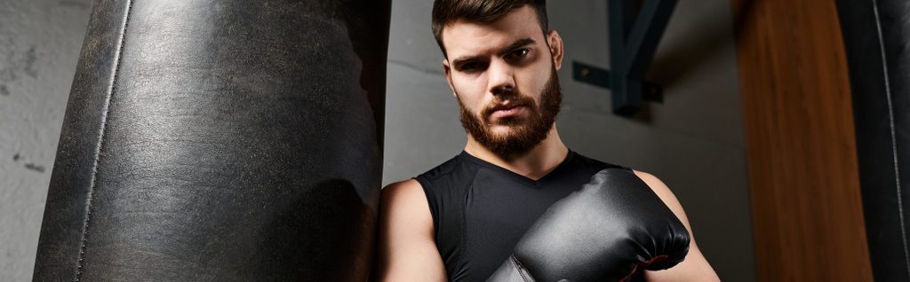 Un bel homme barbu portant des gants de boxe se tient à côté d'un sac de boxe dans une salle de gym, prêt pour une séance d'entraînement. - Photo, image