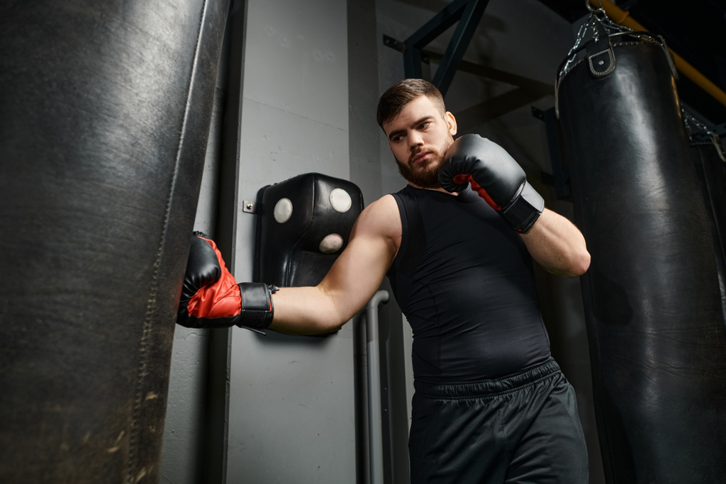 Przystojny mężczyzna w brodzie w czarnej koszuli i czerwonych rękawiczkach bokserskich uderza w worek treningowy na siłowni.. - Zdjęcie, obraz