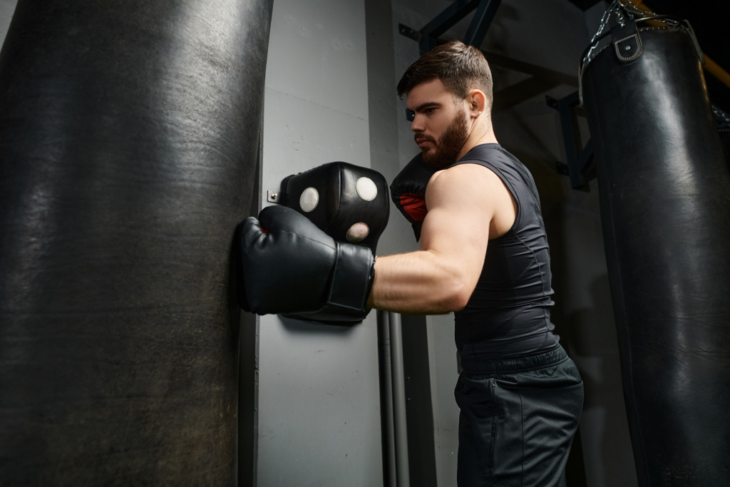 Un hombre guapo con barba, con una camiseta negra y guantes de boxeo, golpea una bolsa en el gimnasio. - Foto, Imagen