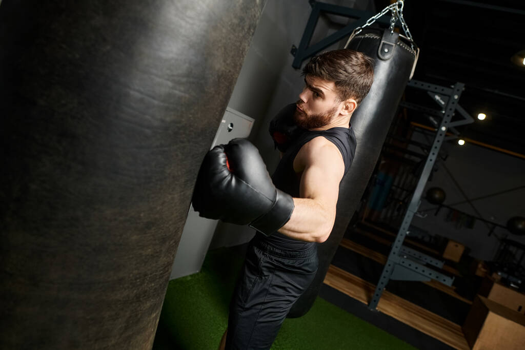 Przystojny mężczyzna z brodą w czarnej koszulce i rękawicami bokserskimi rzucający ciosy w worek treningowy w siłowni. - Zdjęcie, obraz
