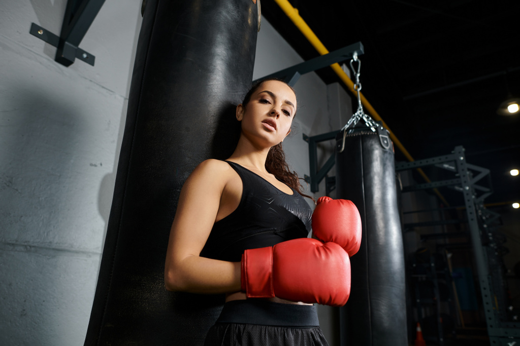 Una sportiva bruna in abbigliamento attivo si trova accanto a un sacco da boxe in una palestra, preparandosi a lanciare un potente pugno. - Foto, immagini