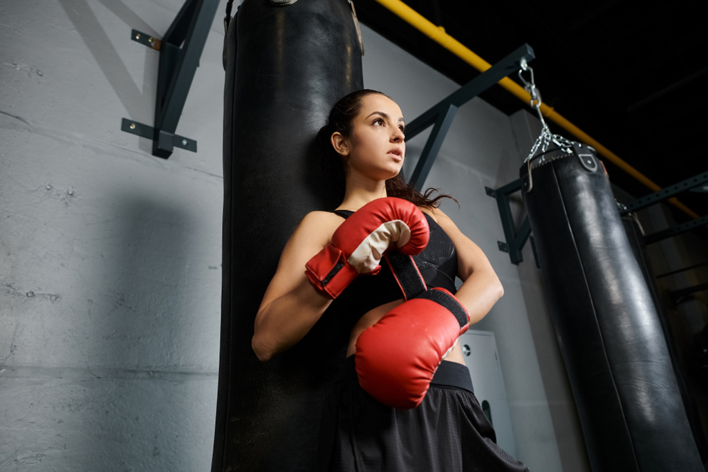 A determined brunette sportswoman wearing red boxing gloves stands confidently next to a punching bag in a gym. - Photo, Image