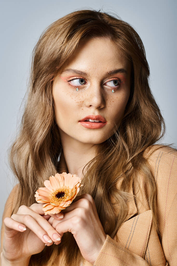 Portret atrakcyjnej kobiety odwracającej wzrok i trzymającej stokrotkę, noszącej makijaż brzoskwiniowy, klejnoty do twarzy - Zdjęcie, obraz