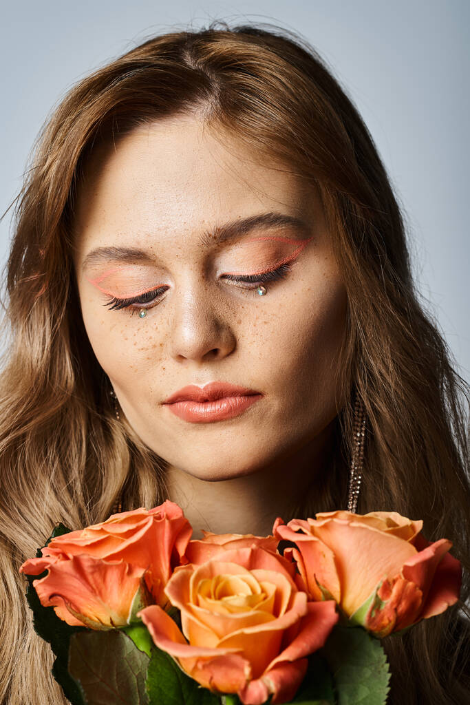 Mulher bonita olhando para rosas e vestindo maquiagem pêssego nu e jóias rosto cintilante - Foto, Imagem