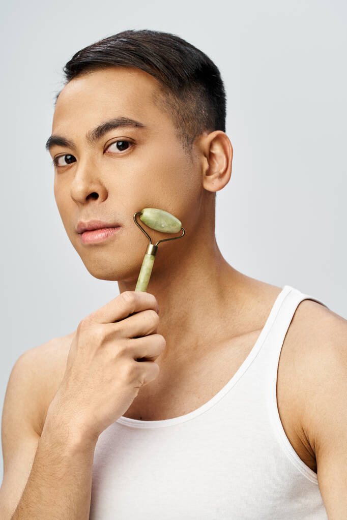 Un bel homme asiatique utilise doucement le rouleau de jade dans le cadre de sa routine de soins de la peau dans un studio gris. - Photo, image