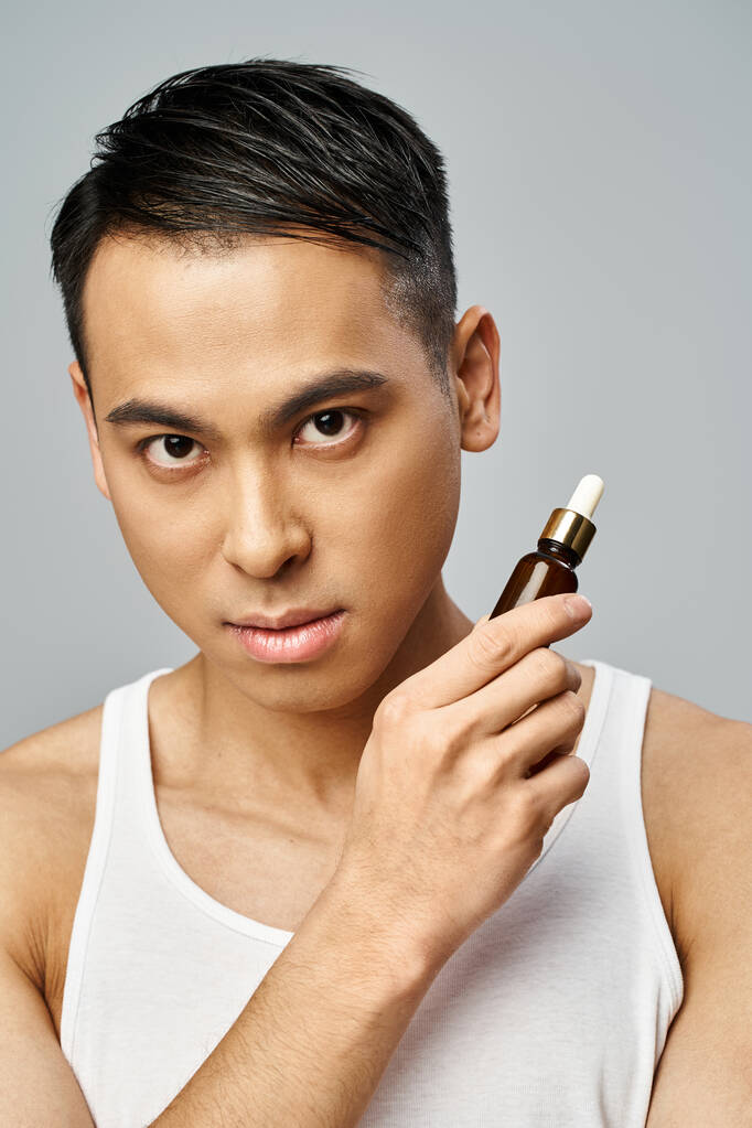 Красивий азіатський чоловік тримає пляшку сироватки в сірій студії, зосереджуючись на рутинному догляді за шкірою. - Фото, зображення