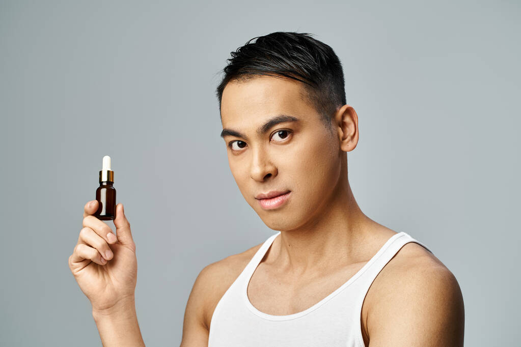 Красивый азиат держит бутылку масла в серой студии, демонстрируя свою рутину по уходу за кожей с точностью и стилем. - Фото, изображение