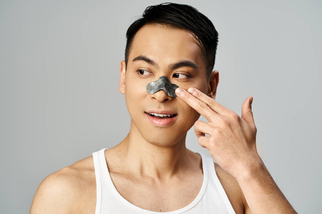 Un hombre asiático con una mascarilla en la mano en la mano hasta la cara en un retrato de rutina de belleza y cuidado de la piel en un estudio gris. - Foto, imagen