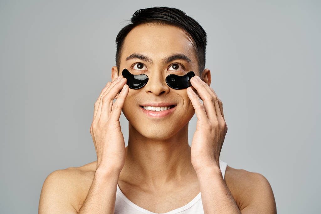 Homem asiático segurando dois círculos negros sobre seus olhos em uma rotina de beleza e cuidados com a pele em um estúdio cinza. - Foto, Imagem