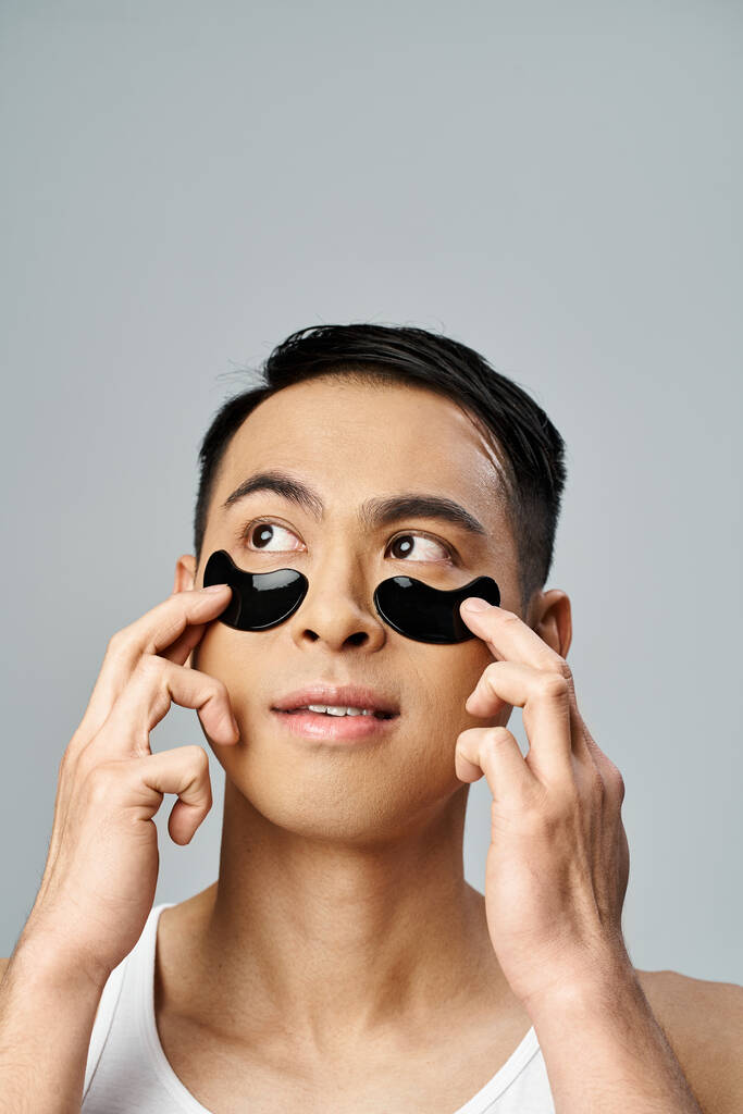 Όμορφος Ασιάτης με μπαλώματα κάτω από τα μάτια που ασχολείται με μια ρουτίνα ομορφιάς και φροντίδας του δέρματος σε ένα γκρίζο στούντιο. - Φωτογραφία, εικόνα