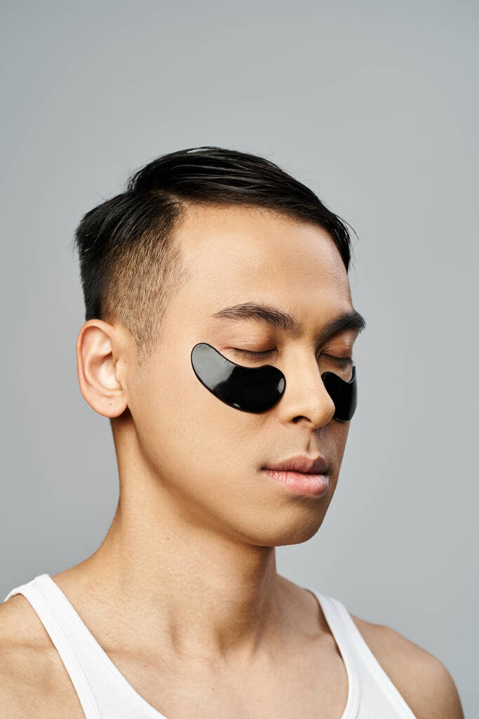 Hezký asijský muž s černým okem páska během krásy rutiny v šedém studiu. - Fotografie, Obrázek