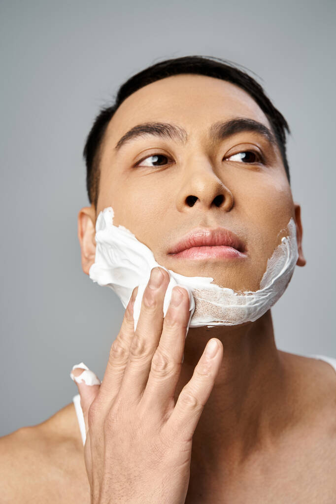 Hombre asiático guapo con espuma de afeitar en la cara en un estudio gris. - Foto, imagen