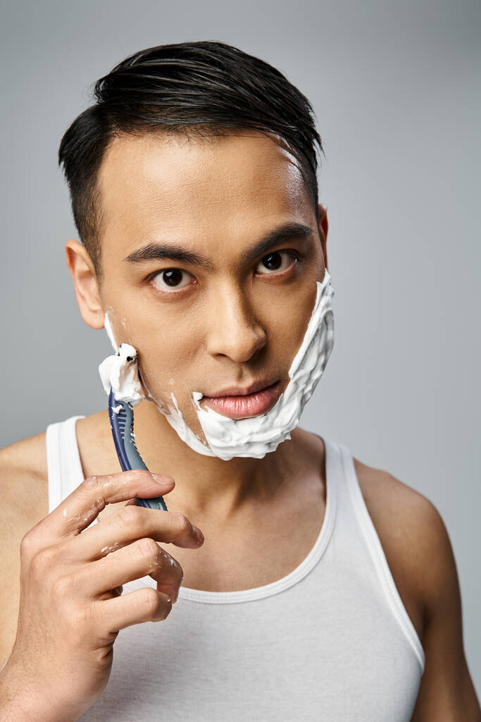 Ein asiatischer Mann mit Rasierschaum im Gesicht rasiert sich aufmerksam mit einem Rasiermesser in einem ruhigen grauen Studio-Ambiente. - Foto, Bild