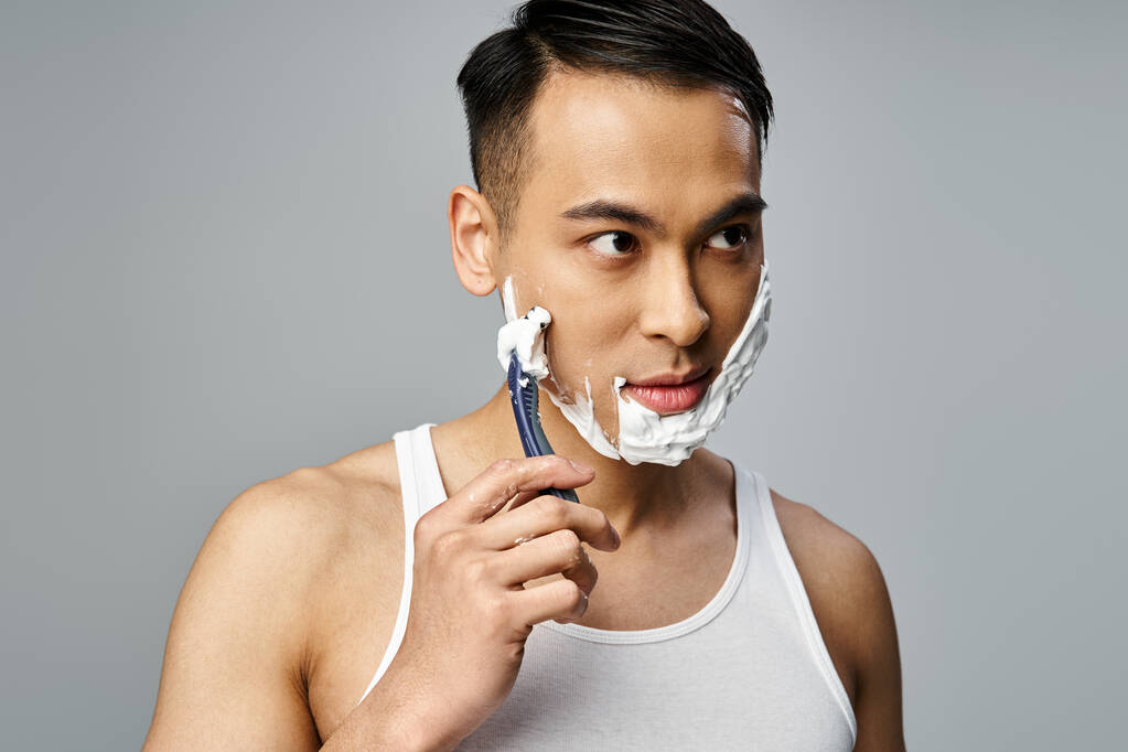 Un retrato de un hombre asiático guapo con espuma de afeitar en la cara, afeitándose cuidadosamente con una navaja en un estudio gris. - Foto, imagen