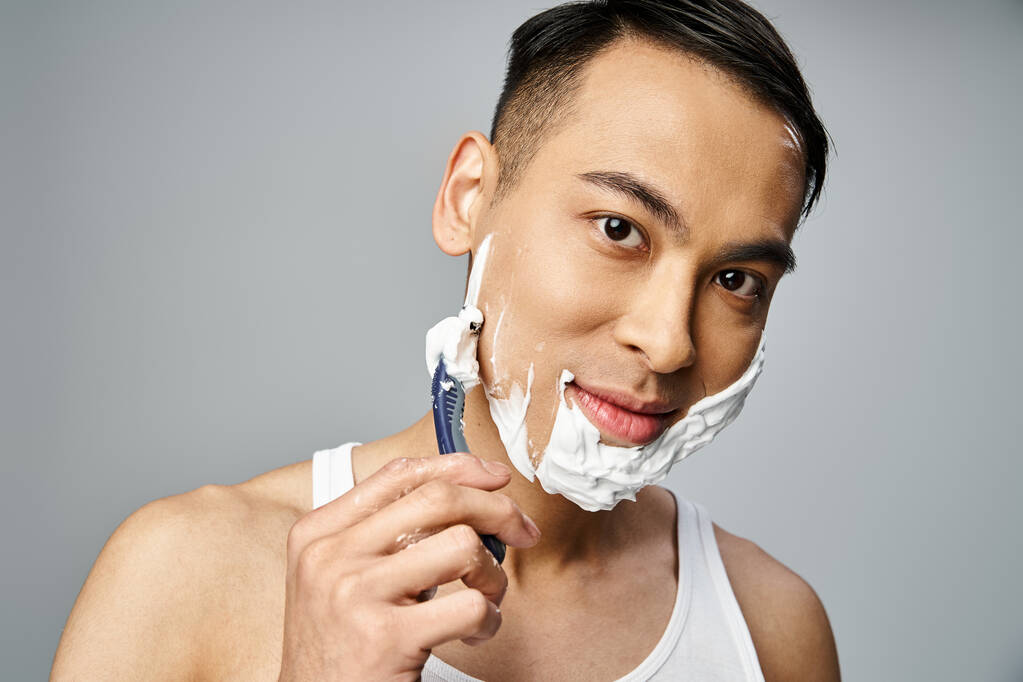 Ein hübscher asiatischer Mann mit Rasierschaum im Gesicht, der in einem grauen Studio ein Rasiermesser benutzt. - Foto, Bild