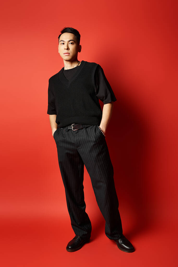 Egy elegáns, jóképű ázsiai férfi áll magabiztosan fekete ruhában, merész vörös háttérrel egy stúdióban.. - Fotó, kép