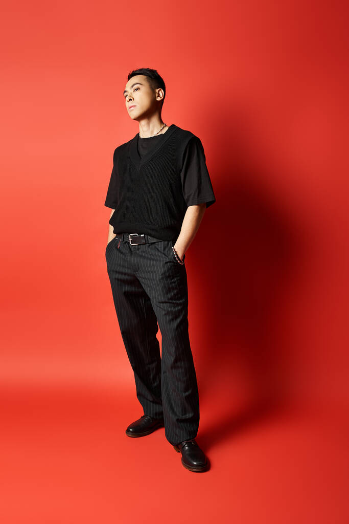 Un Asiatique à la mode en tenue noire se tient en confiance devant un mur rouge frappant dans un décor de studio. - Photo, image