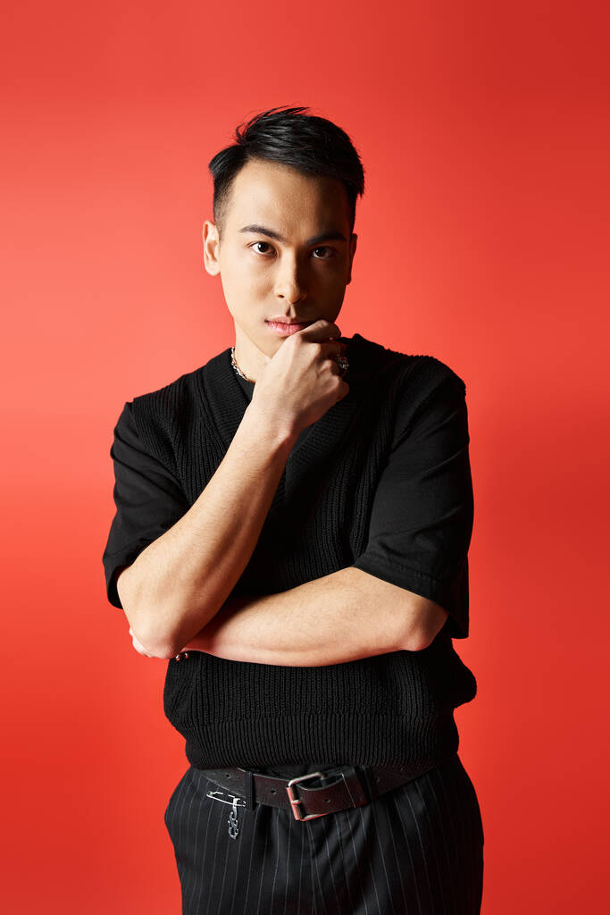 Stylish and handsome Asian man in black attire resting his chin on his hand, deep in thought against a red background. - Photo, Image