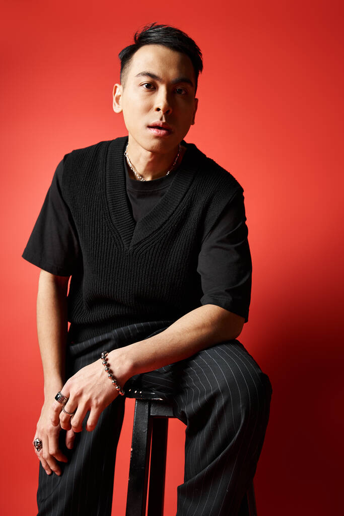 Un uomo asiatico elegante in abito nero si siede su uno sgabello di fronte a un muro rosso sorprendente in un ambiente di studio. - Foto, immagini