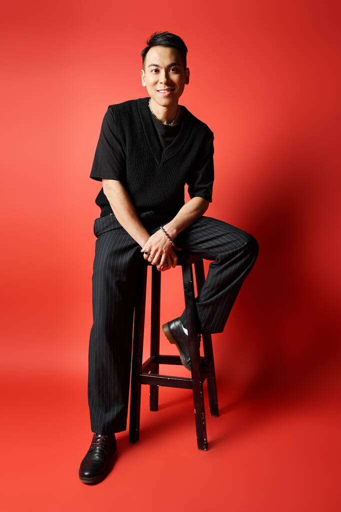 Komea aasialainen mies mustissa vaatteissa istuu tyylikkäästi ulosteella silmiinpistävää punaista taustaa vasten. - Valokuva, kuva