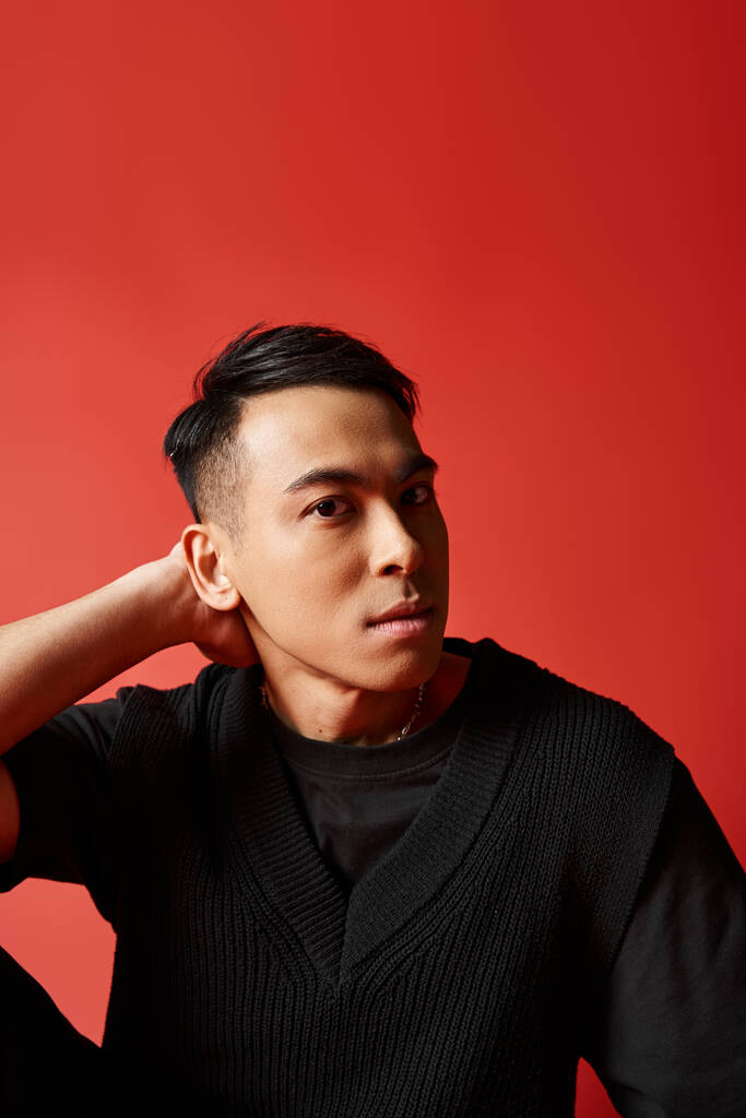 Стильный, красивый азиат в черном свитере позирует на красном фоне в студии. - Фото, изображение