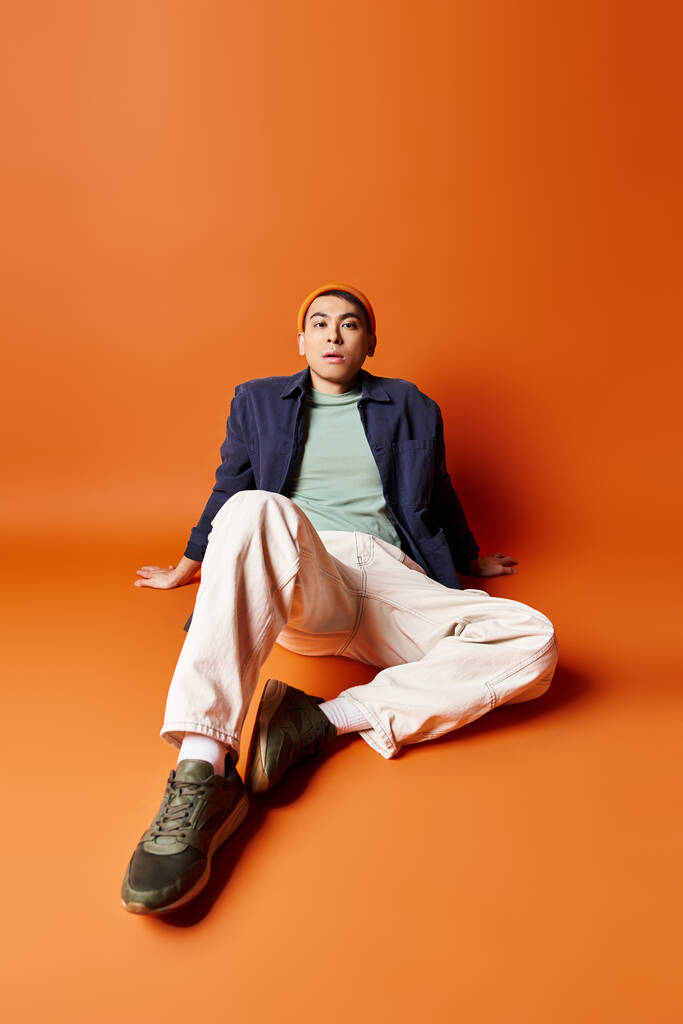 Um homem asiático elegante, bem vestido, senta-se com as pernas cruzadas em um fundo laranja. - Foto, Imagem
