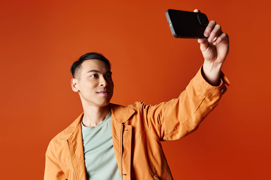 スタイリッシュな服装のハンサムなアジア人男性は,オレンジ色のスタジオの背景に対して携帯電話で写真を撮ります. - 写真・画像