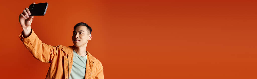 Komea aasialainen mies tyylikkäässä asussa pitäen kännykkää ilmassa oranssia taustaa vasten.. - Valokuva, kuva