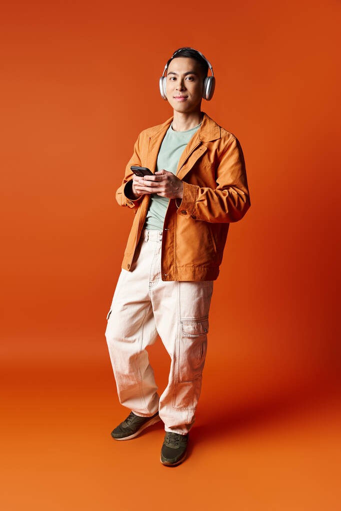 スタイリッシュなアジア人男性は,ヘッドフォンを着用して大胆なオレンジ色の背景の前に立っています. - 写真・画像