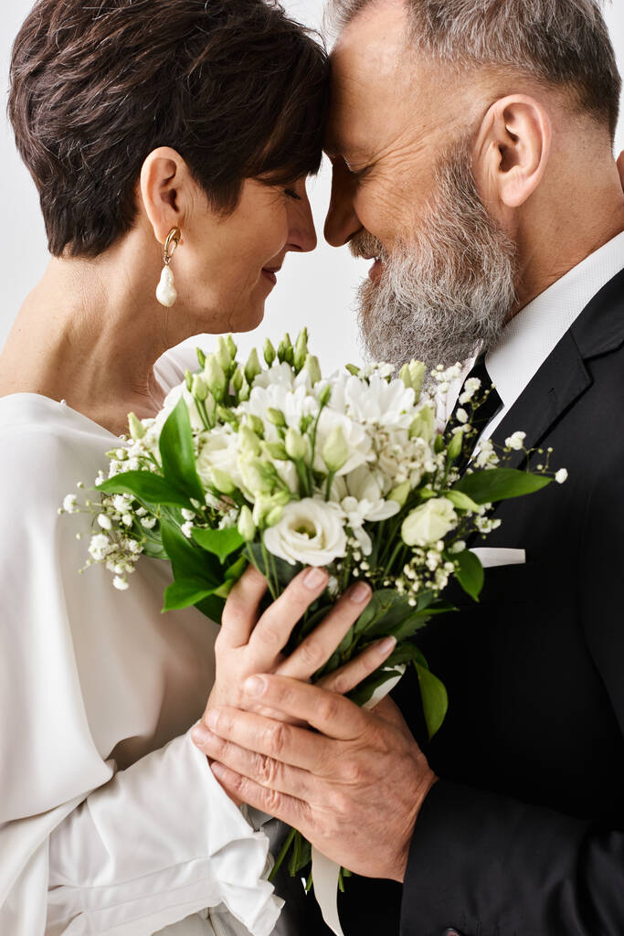 Bruid en bruidegom van middelbare leeftijd in trouwkleding, met een prachtig boeket bloemen, vieren hun speciale dag in een studio setting. - Foto, afbeelding