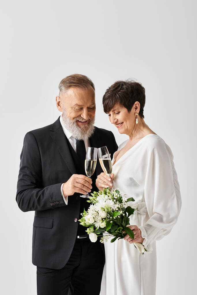 Наречена середнього віку і наречений витончено стоять поруч, тримаючи шампанські окуляри в студійній обстановці, святкуючи свій особливий день. - Фото, зображення