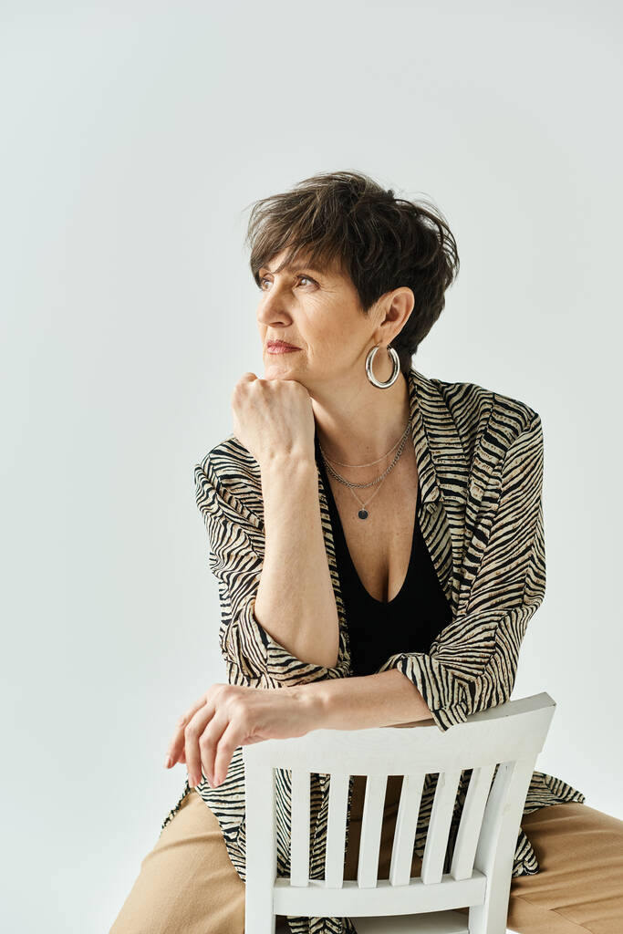 Una mujer de mediana edad con el pelo corto posa elegantemente en la parte superior de una silla blanca en un ambiente elegante estudio. - Foto, imagen