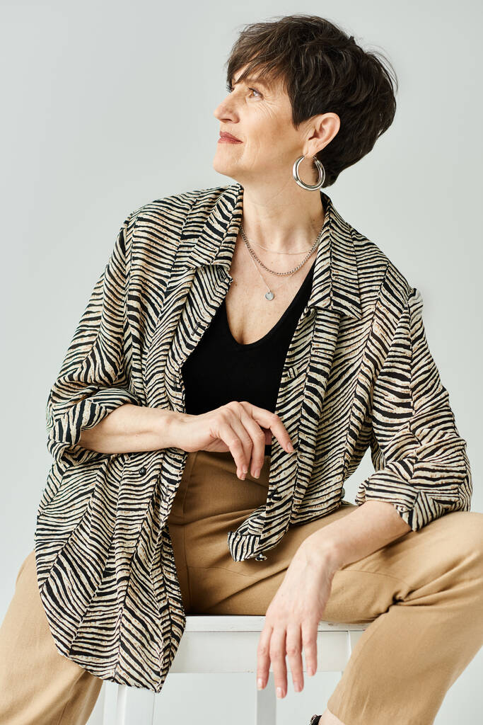 Μια μεσήλικη γυναίκα με κοντά μαλλιά, που φοράει κομψή ενδυμασία, κάθεται με χάρη πάνω από ένα μινιμαλιστικό λευκό σκαμνί σε ένα σκηνικό στούντιο.. - Φωτογραφία, εικόνα