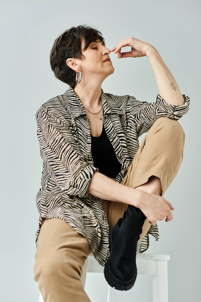 Uma mulher de meia-idade elegante com cabelo curto senta-se graciosamente no topo de um banquinho branco intocado em um estúdio chique. - Foto, Imagem