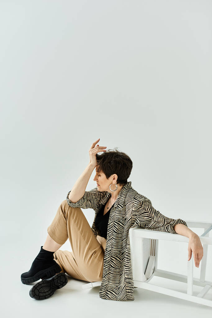 Een vrouw in stijlvolle kleren zit op de grond naast een ongerepte witte stoel in een contemplatieve houding. - Foto, afbeelding