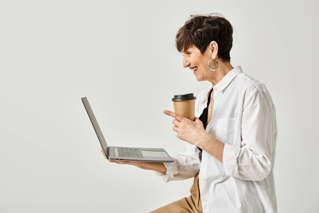 Een vrouw van middelbare leeftijd in stijlvolle kledij die een kop koffie vasthoudt terwijl ze in een studio aan een laptop werkt. - Foto, afbeelding