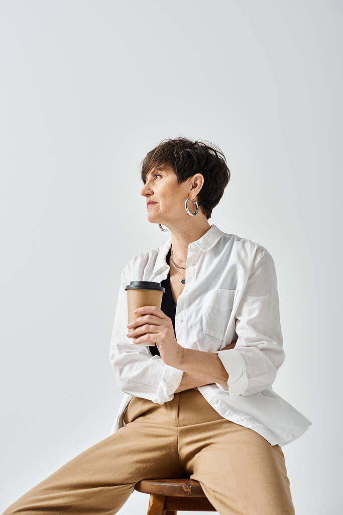 Middelbare vrouw in stijlvolle kleding zit op de kruk met een kopje koffie in een serene studio setting. - Foto, afbeelding