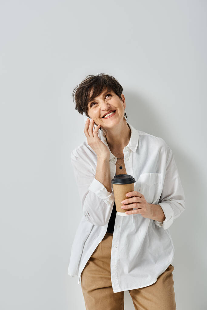 Eine Frau mittleren Alters mit kurzen Haaren, stilvoll gekleidet, Multitasking, indem sie eine Tasse Kaffee in der Hand hält und mit ihrem Handy plaudert. - Foto, Bild