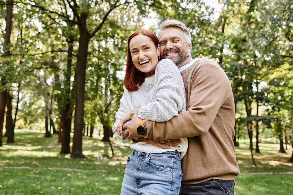 Een man en een vrouw delen een warme knuffel in de serene omgeving van een park. - Foto, afbeelding